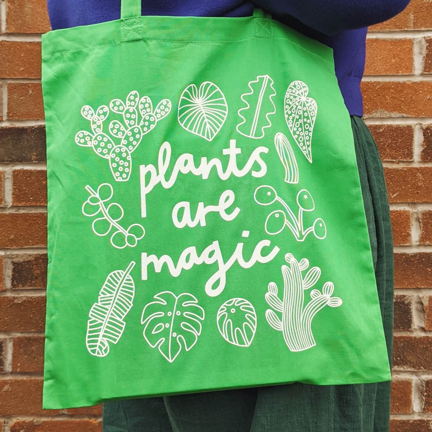 Green tote back with white plants and leaves doodles on and the words &quot;plants are magic&quot; in a playful font. 